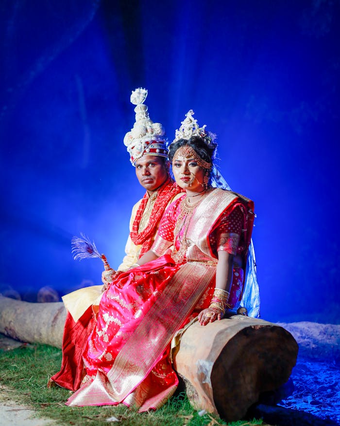 gallery-1 Elegant Bengali couple in traditional wedding attire seated outdoors, under a vibrant blue sky.